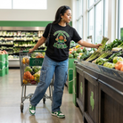 Person shopping in a grocery store with a cart, wearing a black t-shirt with a graphic design "I Create My Own Luck" and "Find A Reason To Smile" on the back.